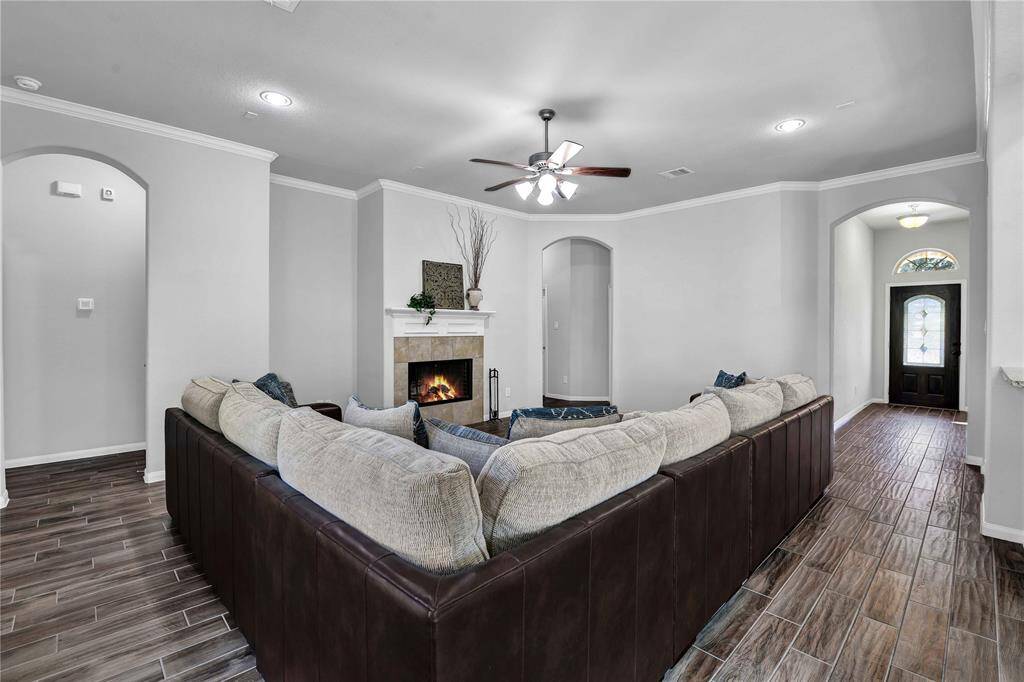 This inviting living room features a cozy corner fireplace, a spacious L-shaped sofa, and elegant wood-style tile flooring. The room is well-lit with recessed lighting and a ceiling fan, creating a warm, welcoming atmosphere. Arched doorways lead to an open hallway and the front entrance, enhancing the home's flow and charm.
