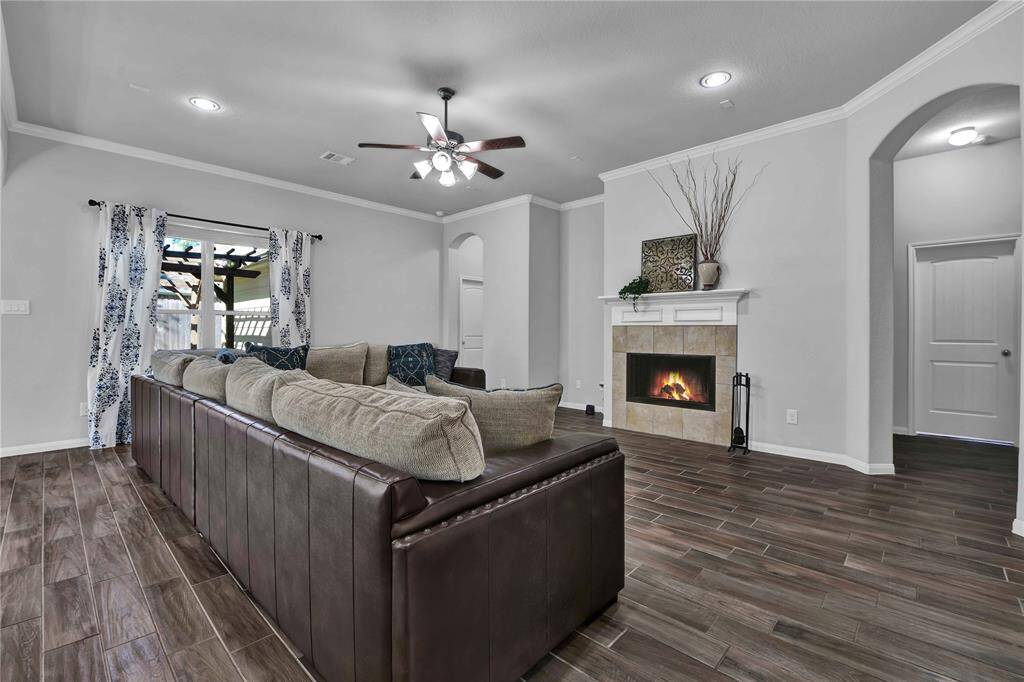 This spacious living room features a cozy fireplace with a tiled surround, elegant wood-look flooring, and a stylish ceiling fan. Large windows add natural light, complemented by tasteful drapery. The area is perfect for relaxation and entertaining.