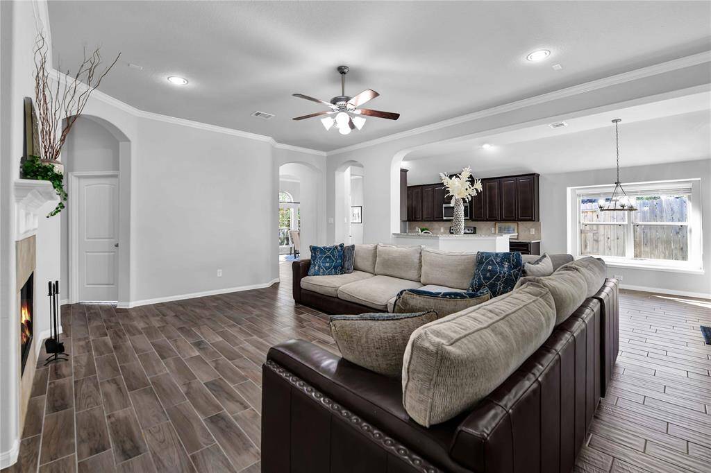 This spacious living area features an open floor plan with wood-look tile flooring, a cozy fireplace, and a large sectional sofa. The modern kitchen is visible, boasting dark cabinetry and stainless steel appliances. A ceiling fan and recessed lighting add to the bright, airy feel, complemented by a large window for natural light.