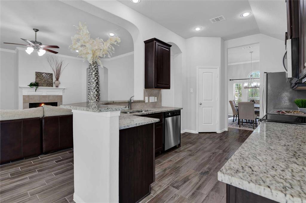 This home features an open-concept kitchen and living area with sleek granite countertops, dark wood cabinetry, and stainless steel appliances. The space flows into a cozy living room with a fireplace, and a dining area with large windows offering natural light. The neutral color palette and modern fixtures create an inviting atmosphere.