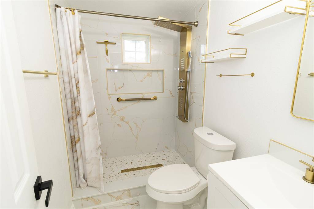 This is a modern and sleek full bathroom featuring white fixtures, marble flooring, and gold accent hardware.