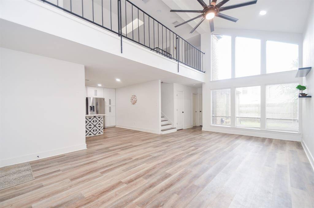 This is a spacious, modern living area with high ceilings, large windows allowing for ample natural light, a contemporary ceiling fan, and a stylish open staircase leading to an upper level. The room features light wood flooring and a neutral color palette.