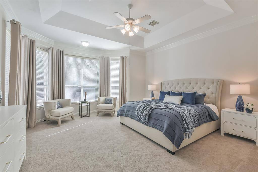 The primary bedroom has carpeted floors, double crown molding, a tray ceiling, a ceiling fan, custom drapes, and a bay window with 2” blinds.