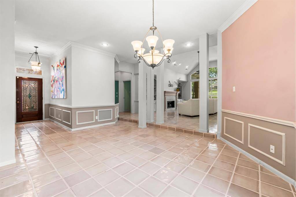 The photo shows a spacious, well-lit entryway with tile flooring, leading into a living area with a fireplace. The space features elegant wainscoting, neutral wall colors, and a mix of modern and traditional light fixtures.