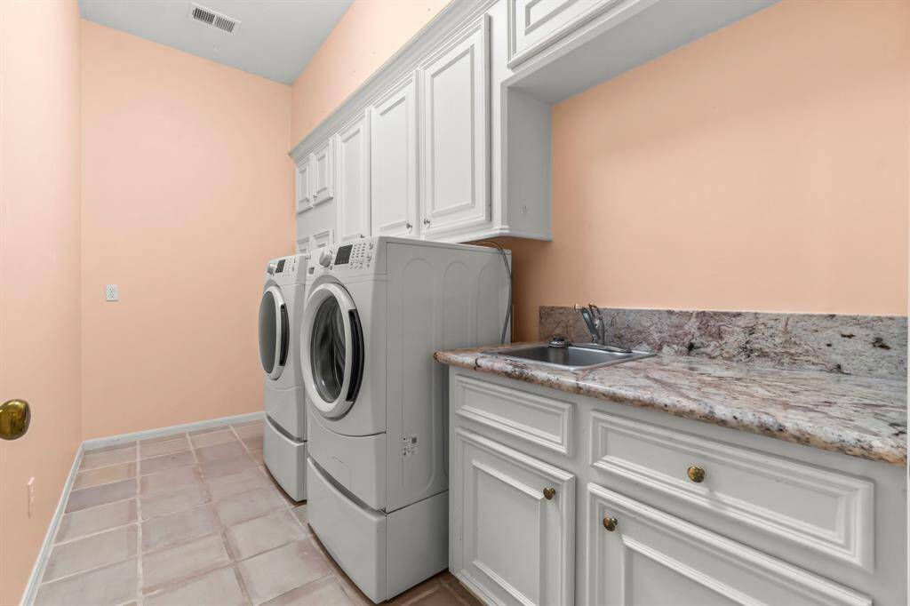 This is a dedicated laundry room with a side-by-side washer and dryer, ample white cabinetry for storage, and a sink with a granite countertop. The room has tile flooring and is painted in a light peach/pink hue.