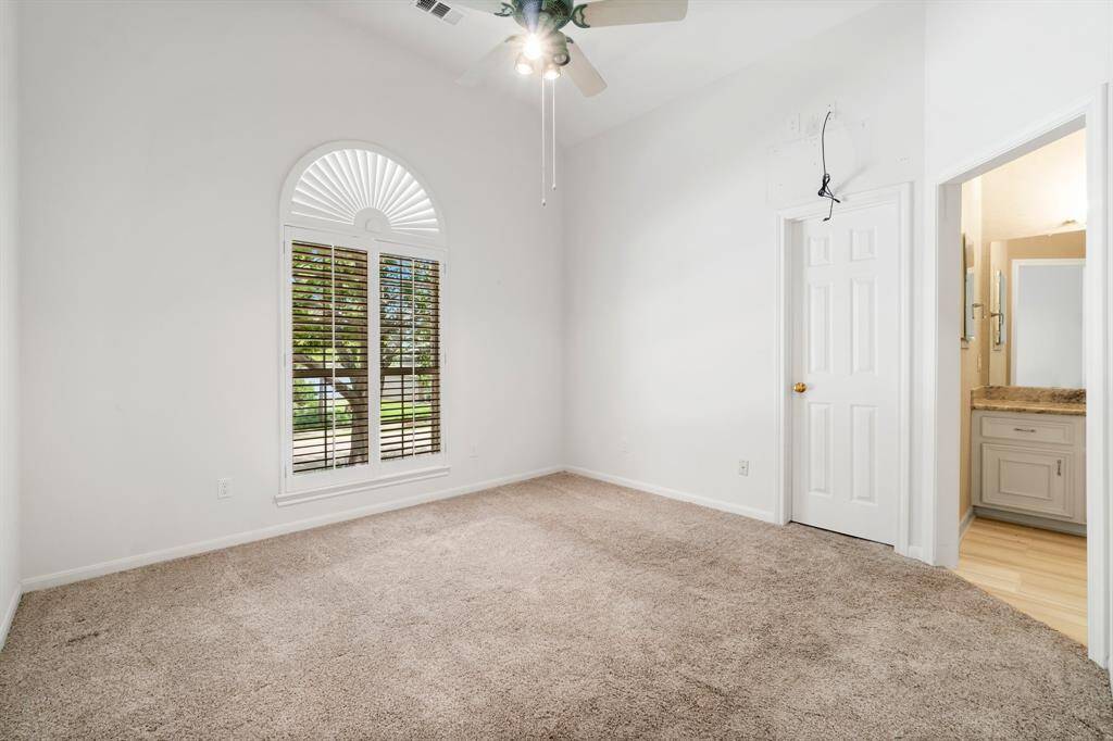 This is a spacious room with high ceilings, large arched window with blinds, plush carpeting, and a ceiling fan. There is also a doorway leading to another room with a partial view of a bathroom vanity.