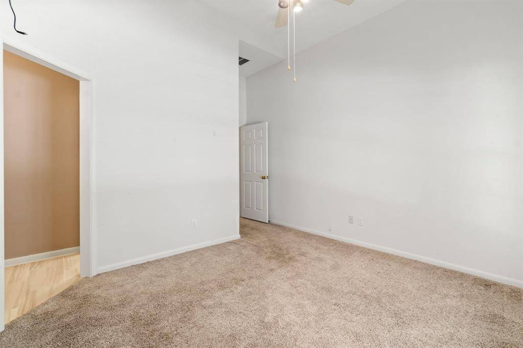 This is a spacious room with neutral walls and carpet flooring, featuring a high ceiling with a fan and good natural light potential. There's an open doorway leading to another room with hardwood floors and a closed white door, suggesting a versatile space that could be personalized.