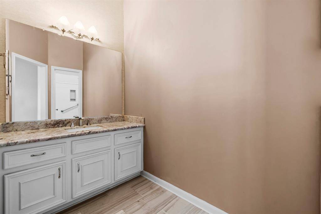 This is a bathroom featuring a white vanity with a granite countertop, under-mount sink, and a large mirror, complemented by overhead lighting. Neutral wall colors and wood-look flooring give the space a warm, modern feel.