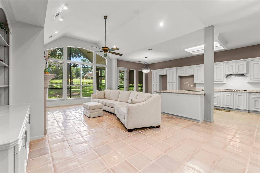 This is a bright, open-concept living space featuring a high vaulted ceiling with large windows overlooking a green outdoor area. The room seamlessly transitions into a modern kitchen with white cabinetry and a central island. The area is finished with tiled flooring, providing a spacious and airy feel.