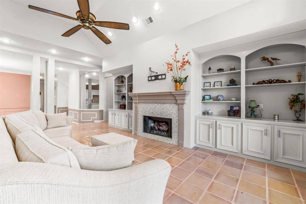 This is a spacious, well-lit living space featuring a cozy fireplace with a decorative mantel, built-in shelving for storage and display, and a ceiling fan for comfort. The room has an open layout with easy access to adjoining rooms, highlighted by  tiled flooring and neutral color walls.
