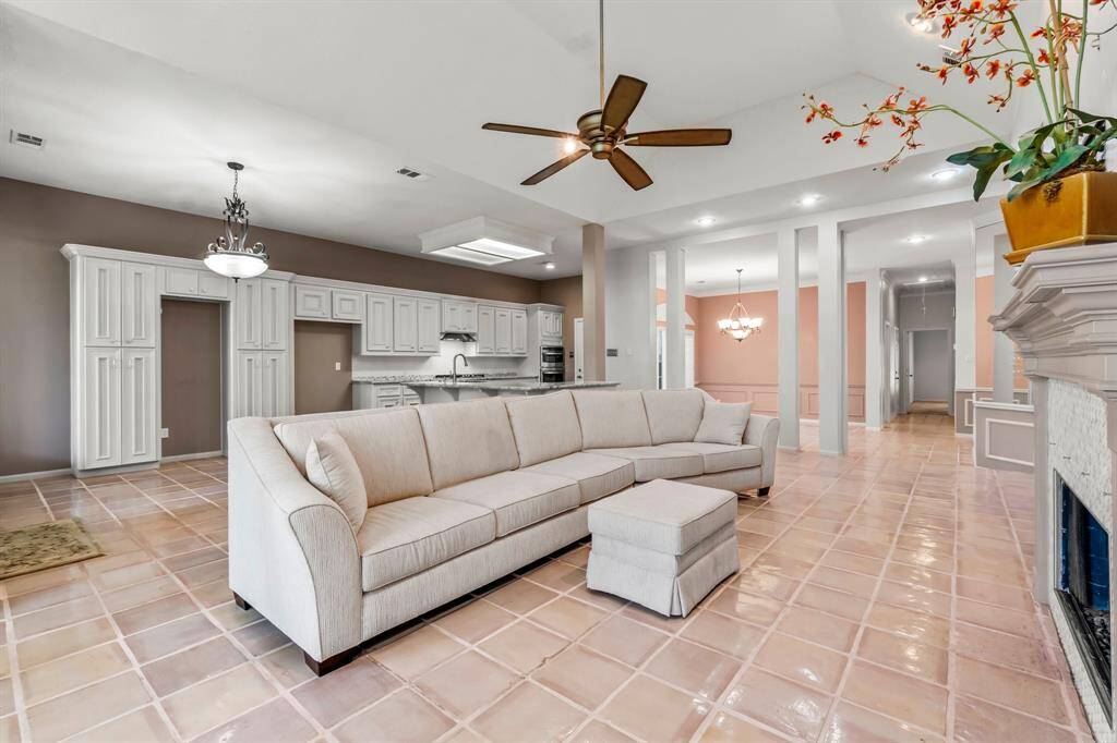 This is a spacious, open-concept living area with tile flooring, featuring a large sectional sofa and a cozy fireplace. The room flows seamlessly into the kitchen with white cabinetry and is illuminated by natural light and a stylish ceiling fan. Elegant columns and warm color accents create a welcoming ambiance.