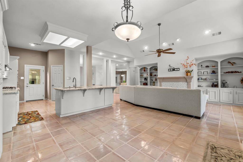 This is a spacious, open-concept living area featuring high ceilings with recessed lighting, a ceiling fan, a fireplace with built-in shelving, and a large kitchen island. The room has tile flooring and neutral color tones.