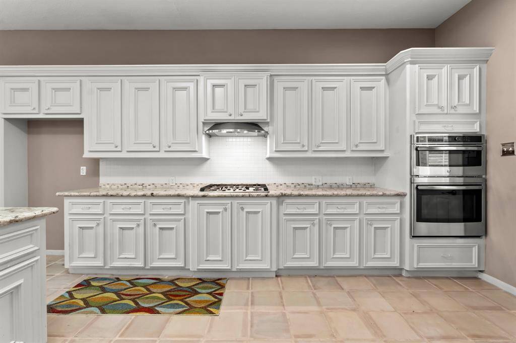 This kitchen features white cabinetry, granite countertops, stainless steel appliances, including a double wall oven and gas cooktop, with a tile backsplash and tile flooring. It has a neutral color palette and ample storage space.