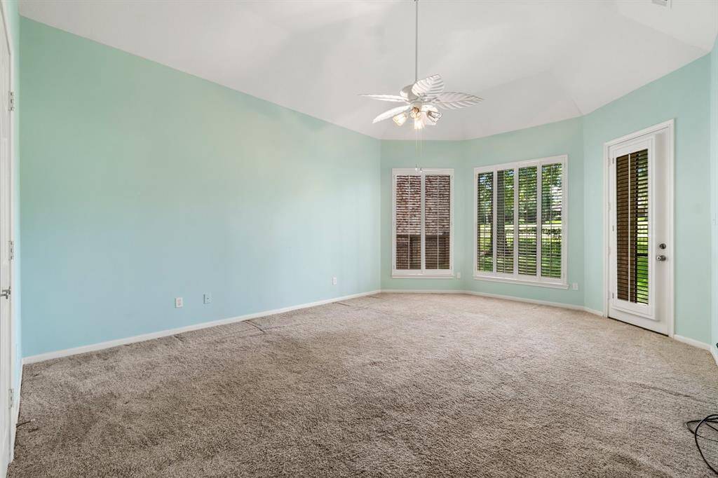 This is a spacious, well-lit room featuring light green walls, beige carpet flooring, a ceiling fan, and ample natural light from windows with plantation shutters and a glass door leading outside.