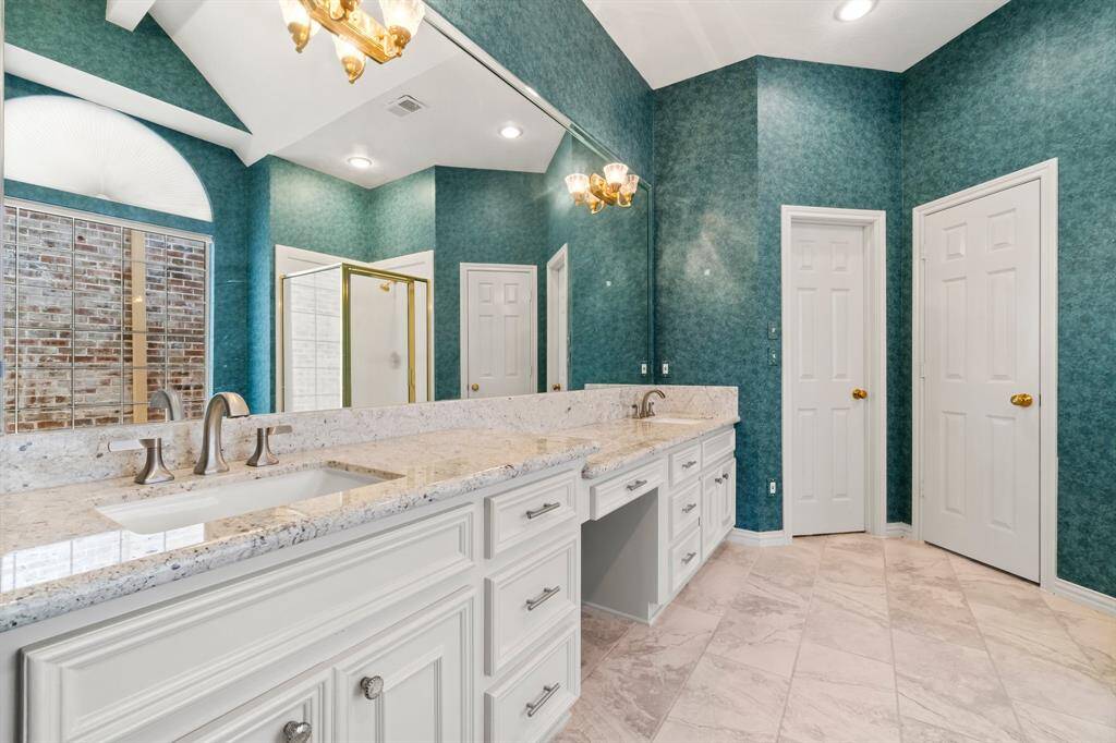 This is a spacious bathroom featuring a double vanity with granite countertops, white cabinetry, and a full-width mirror. It has a tiled walk-in shower, a separate toilet room, and elegant tiled flooring. The room is finished with textured teal wallpaper and has ample lighting.
