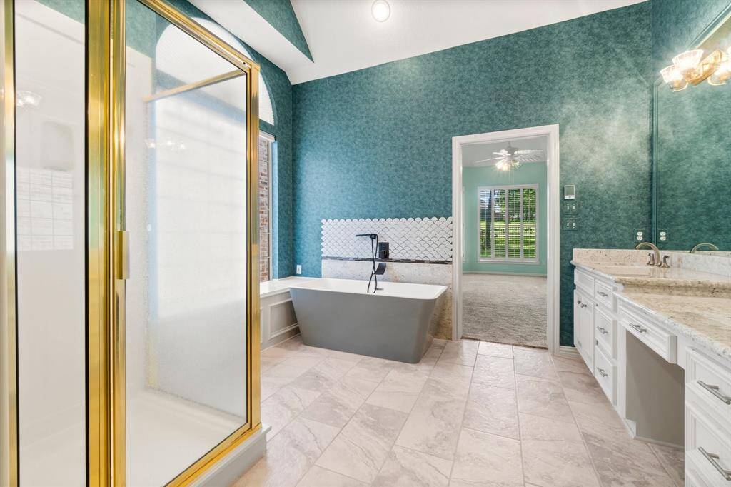This is a spacious, well-lit bathroom featuring a modern freestanding tub, a separate enclosed shower, dual vanities with ample counter space, and a striking blue-patterned wallpaper. A window provides natural light and a view to the outside.