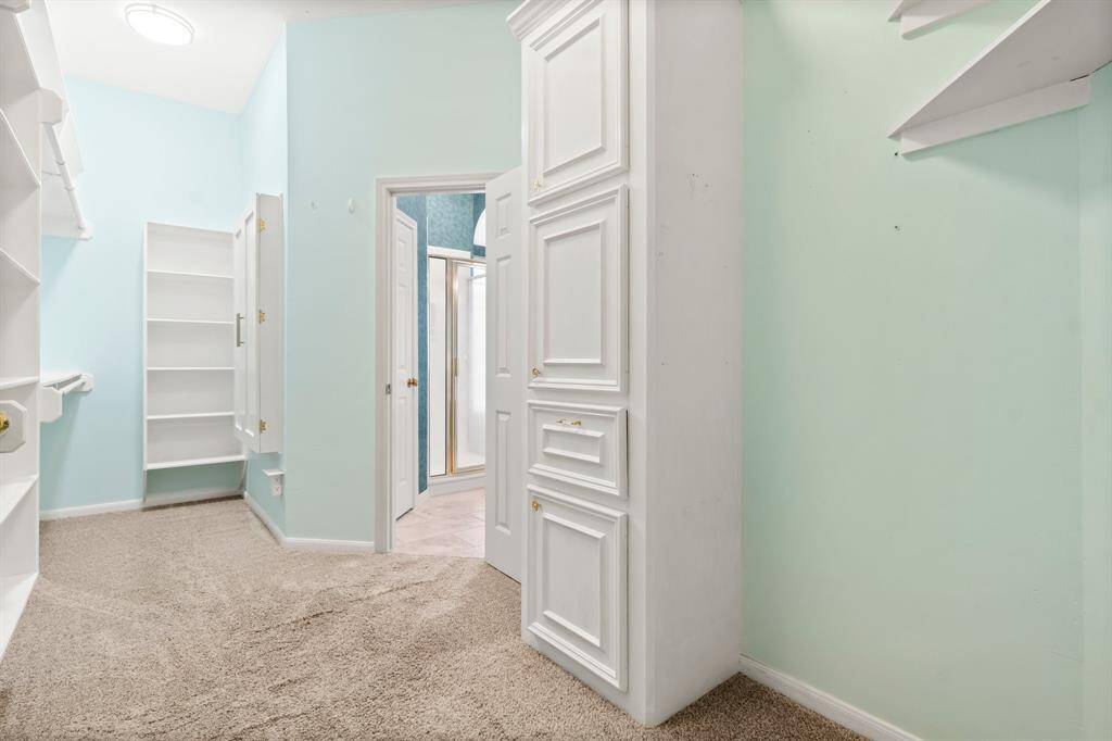Primary bedroom closet with light blue walls, beige carpeting, built-in white shelving, and two doors leading to other areas. It has a bright and airy feel, perfect for a home office or guest room.