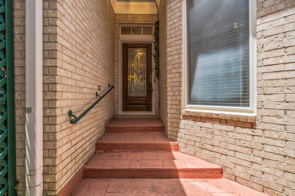 This is a covered entryway featuring terracotta-colored steps leading up to a decorative glass front door, flanked by brick walls, with a large window to one side. There is a green metal handrail for safety.