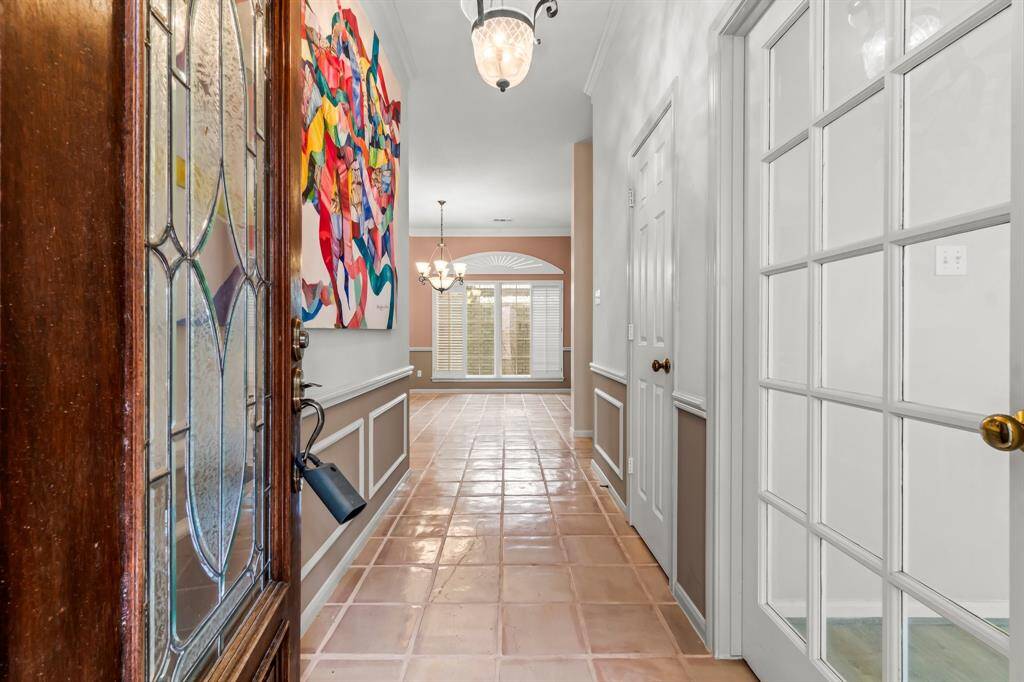 This is a bright and welcoming hallway featuring a tiled floor, wainscoting, and neutral wall colors. The space is enhanced by natural light from a window at the end and includes a decorative glass-paneled door, adding an elegant touch.