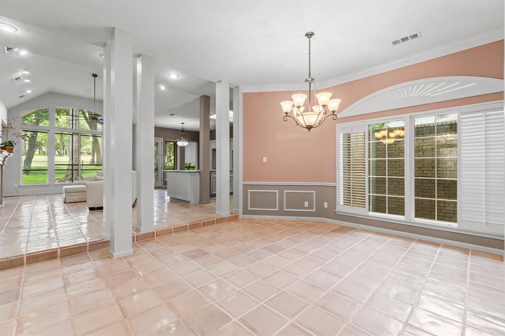 This is a spacious and bright room featuring glossy tile flooring, elegant white columns, and ample natural light from large windows with plantation shutters. The room offers a view of the greenery outside and includes decorative chair rail molding and a stylish chandelier, providing a blend of classic and contemporary design elements.