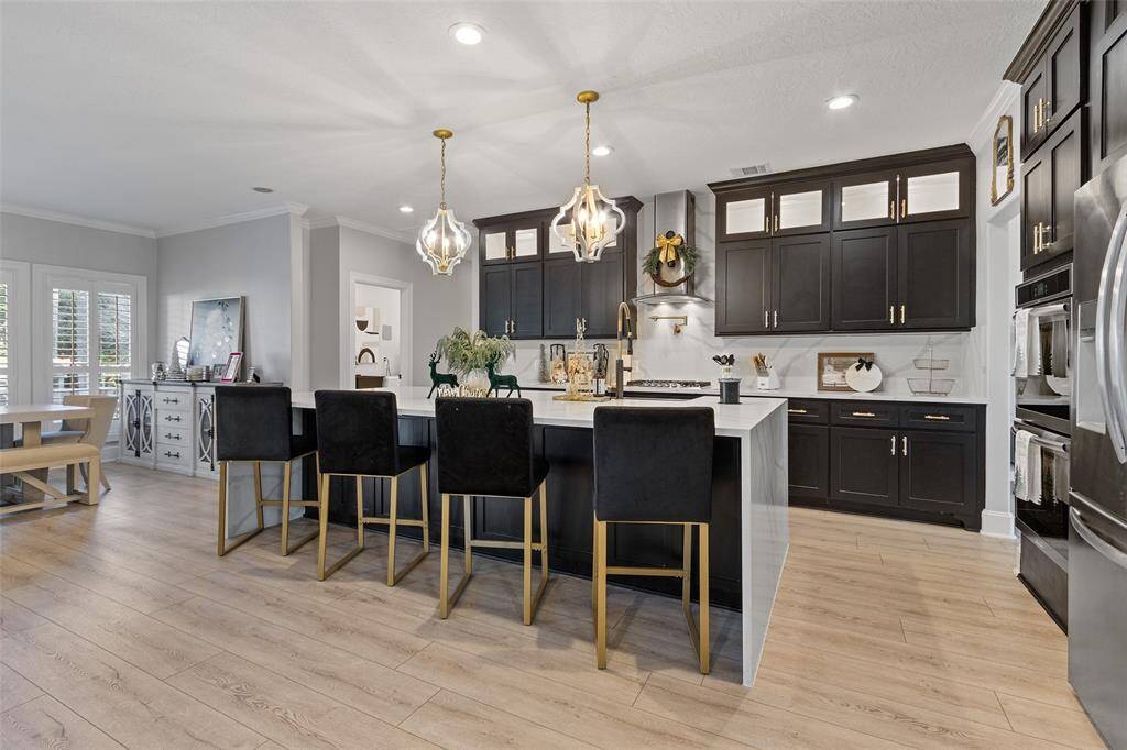 This kitchen showcases a modern kitchen with plenty of cabinets and a large island featuring stylish pendant lighting. It includes a dining area with natural light, wood flooring, and sleek appliances, creating a warm and inviting atmosphere. Cabinets have been painted a tan color recently.