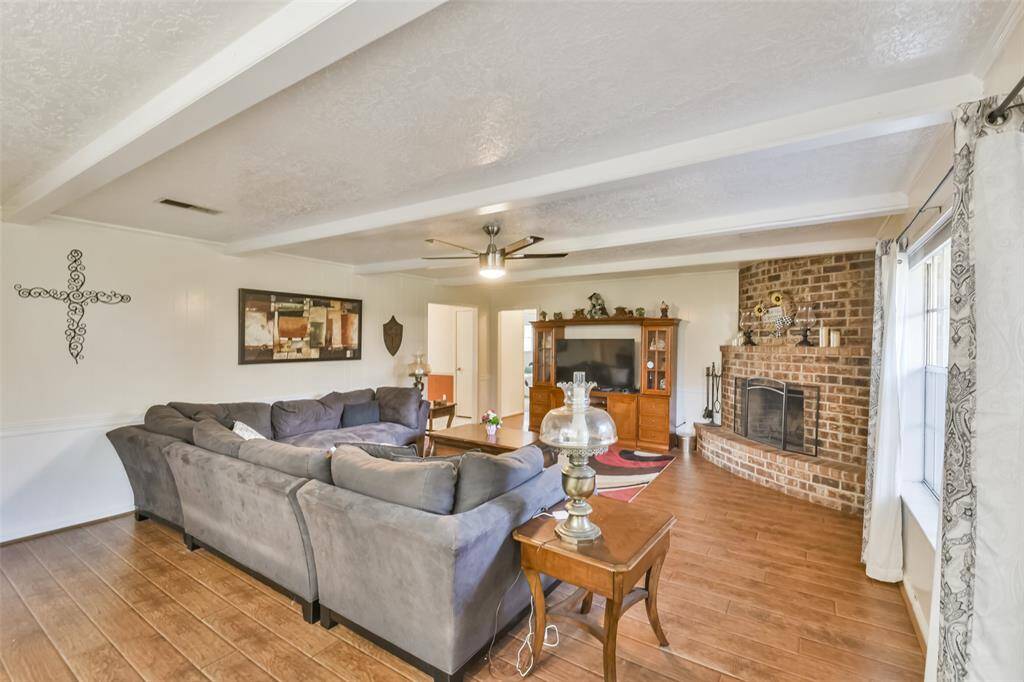 Spacious living room featuring a cozy brick fireplace, wood-look flooring, and a large sectional for ample seating. The space is accented by a ceiling fan, natural light from expansive windows, and an enterainment center for added functionality