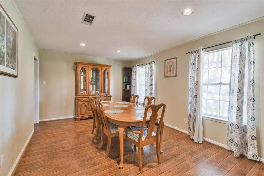 Bright and spacious dining room featuring a classic wood dining set and matching china cabinet. The wood-look flooring complements the neutral walls, while large windows with patterned curtains provide an abundance of natural light for an inviting atmosphere