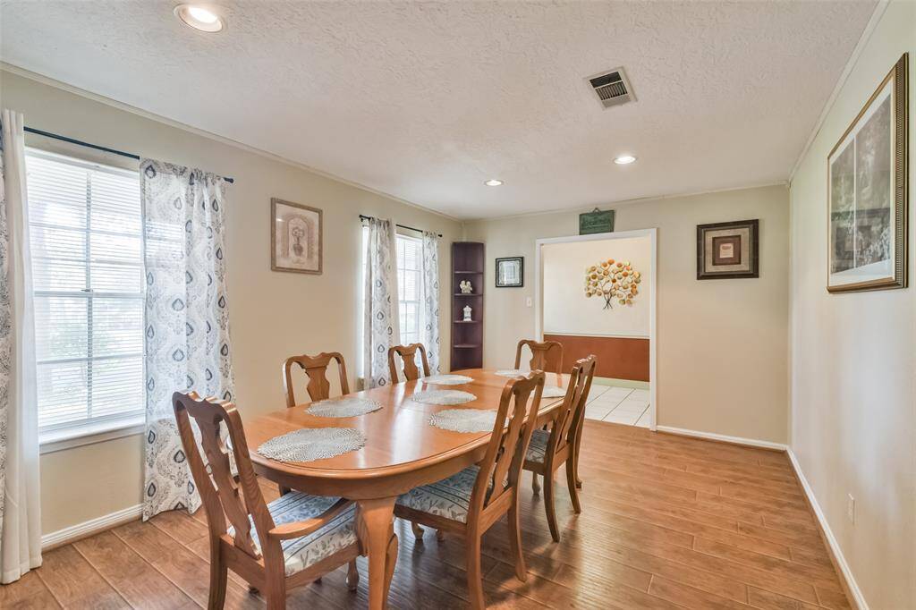 Bright and airy dining room featuring a large wooden table, accented by patterned curtains on large windows. The open design connects seamlessly to an adjacent room, while recessed lighting and warm wood-look flooring enhance the inviting atmosphere