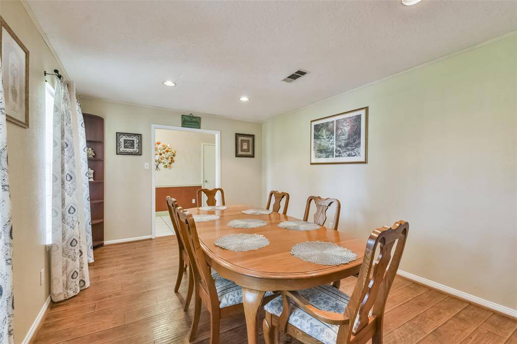 Charming dining room featuring a polished wood table with seating for eight, complemented by wood-look flooring and soft neutral walls. Recessed lighting and large windows with patterned curtains create a bright and welcoming space, perfect for entertaining