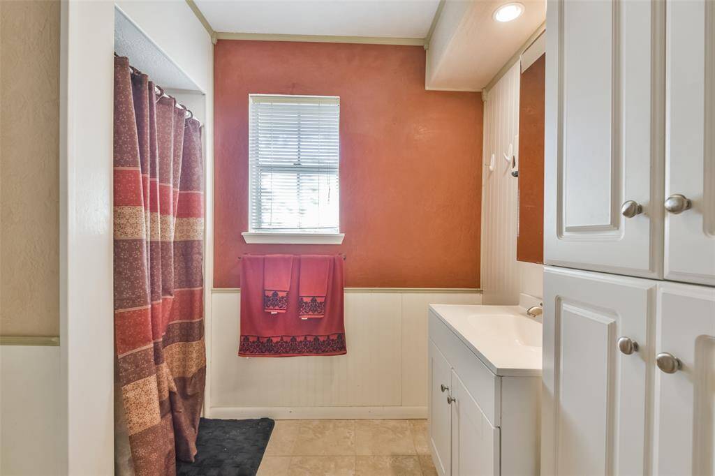 Cozy primary bathroom featuring warm terracotta walls with beadboard wainscoting, a window for natural light, and a single vanity with white cabinetry. The space includes a walk-in shower with a patterned curtain and built-in storage for convenience and organization