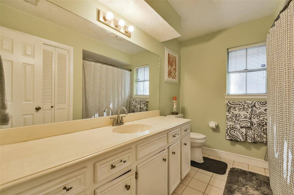 Bright second bathroom featuring a long vanity with ample counter space, a large mirror, and stylish lighting. The soft green walls add a soothing touch, while the shower-tub combo with a patterned curtain and a window provide both functionality and natural light
