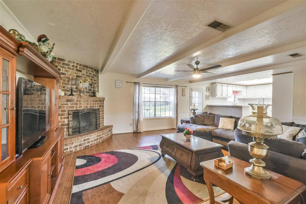 Spacious living room with a warm brick fireplace as the focal point, complemented by a beamed ceiling and large window offering natural light. Open layout flows seamlessly into the kitchen, creating an inviting atmosphere for relaxation and entertaining