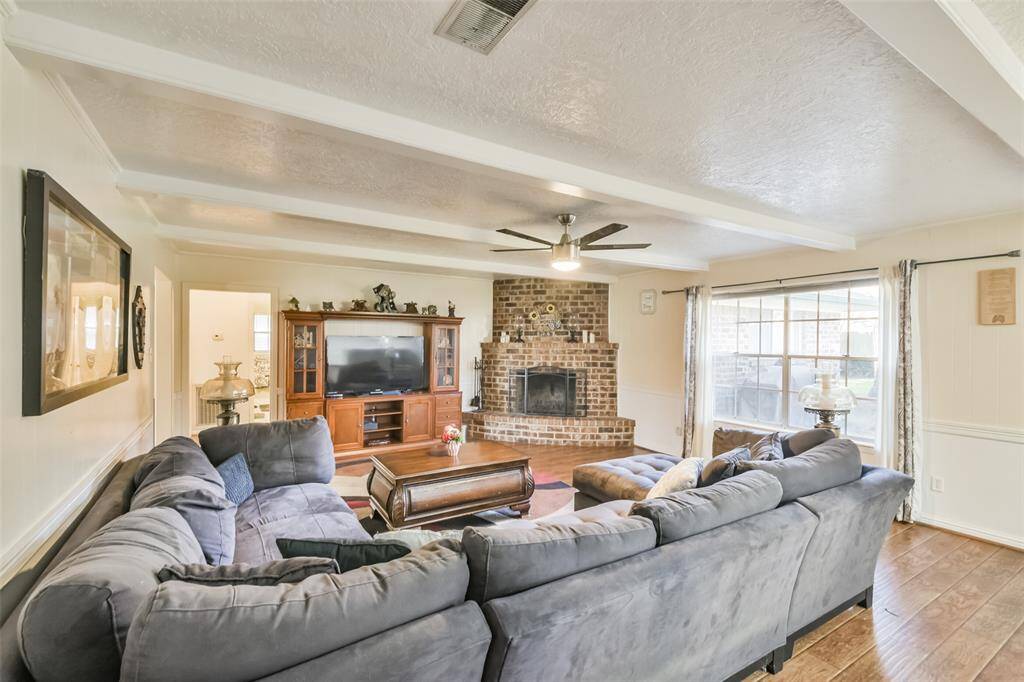 Inviting living room showcasing a warm brick fireplace, a large sectional sofa, and wood-look flooring. The open layout is enhanced by a ceiling fan, natural light from a picture window, and a media center for added storage and entertainment