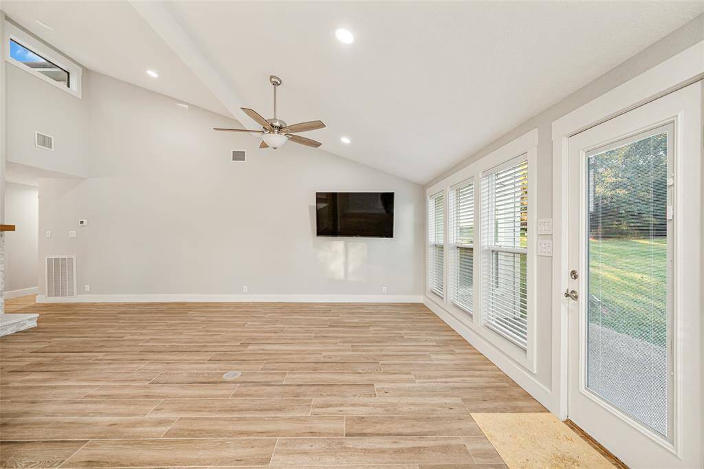 Bright, spacious living room with high ceilings and wood-like flooring. Features large windows offering plenty of natural light, a ceiling fan, and a wall-mounted TV. French doors lead to a lush outdoor view.