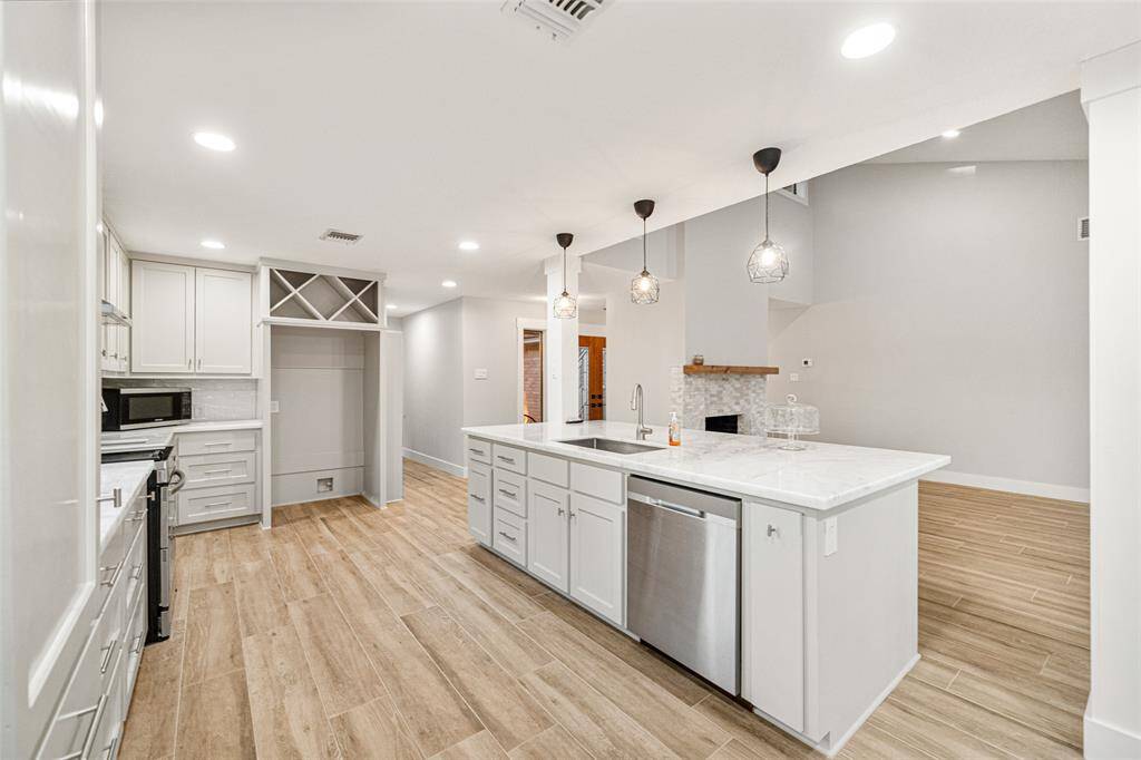 This photo showcases a modern, open-concept kitchen with white cabinetry, a large island, and stainless steel appliances. The space features pendant lighting, hardwood-style flooring, and an adjacent living area with a fireplace.