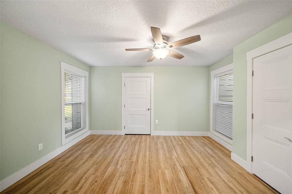 Bright room with light green walls, wood flooring, and ample natural light from two large windows. It features a ceiling fan and a white door, offering a cozy and inviting atmosphere.