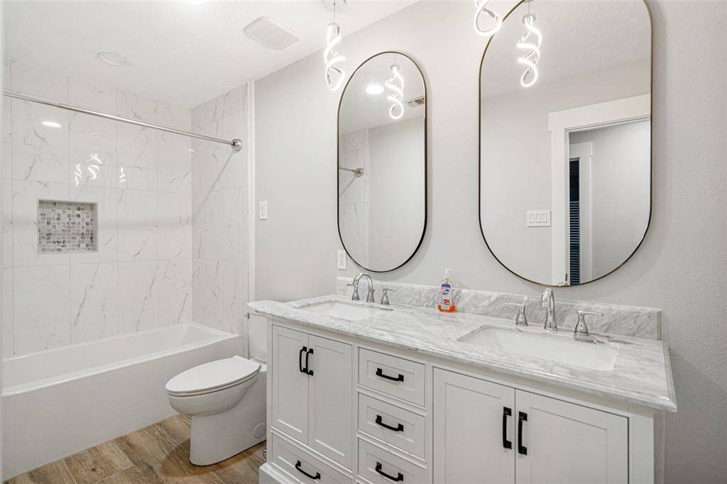 This bathroom features a modern design with a double vanity topped with , stylish oval mirrors, and sleek lighting. The shower-tub combo is accented with elegant white tile and a built-in niche.