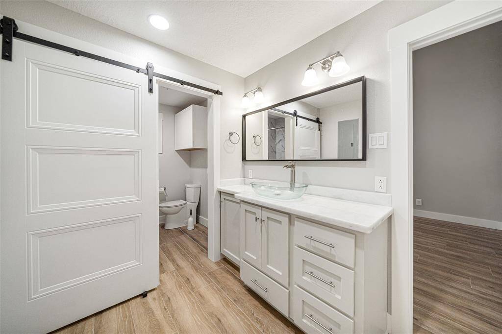 This photo showcases a modern bathroom featuring a sleek double vanity with a glass vessel sink, stylish lighting, and a large mirror. It includes a sliding barn door leading to a toilet area, all set on wood-style flooring.