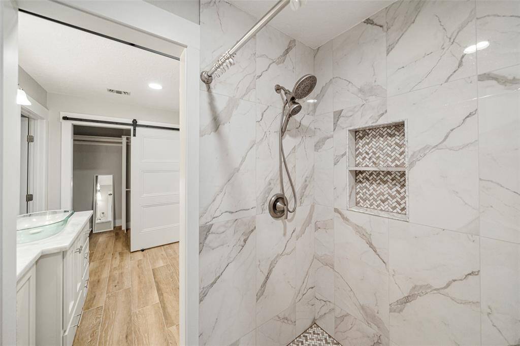 This photo showcases a modern bathroom with a sleek walk-in shower featuring marble-patterned tiles and built-in shelving. Adjacent is a vanity with a glass sink, leading to a spacious closet with a sliding barn door. The wood-look flooring adds warmth to the space.