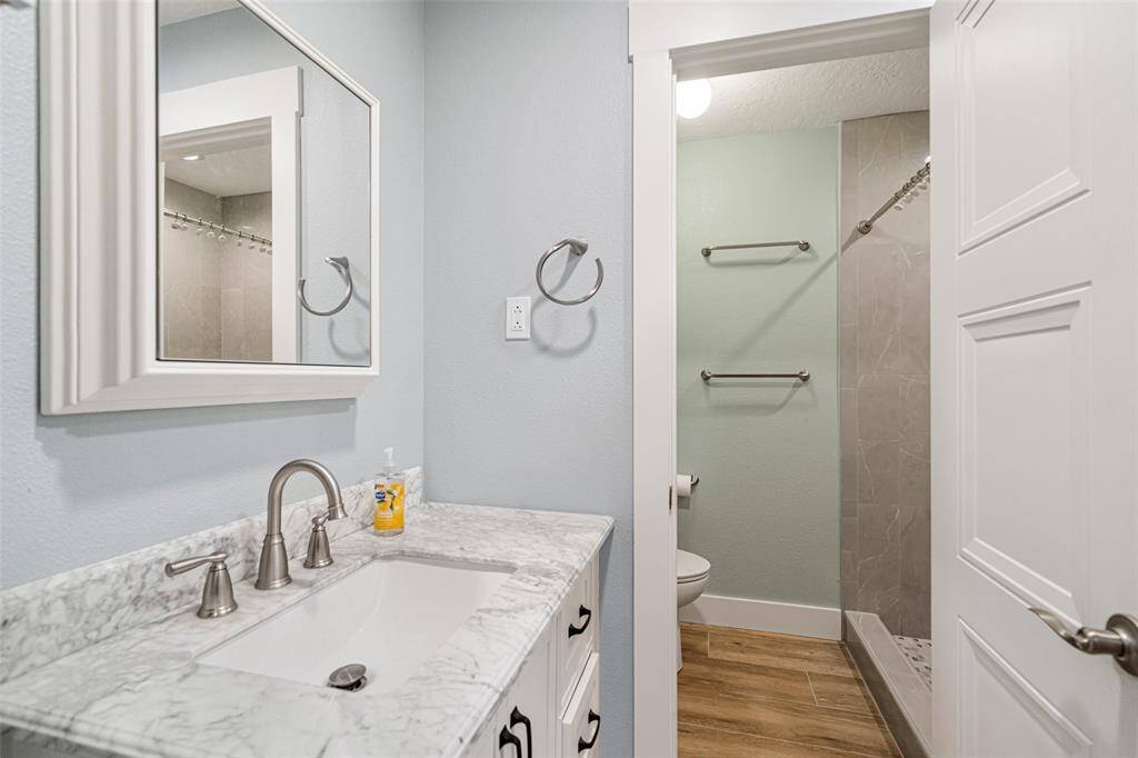 This bathroom features a modern design with a granite countertop, sleek faucet, and a large mirror above the vanity. It has a walk-in shower with tiled walls and a wooden-style floor, creating a clean and inviting space.