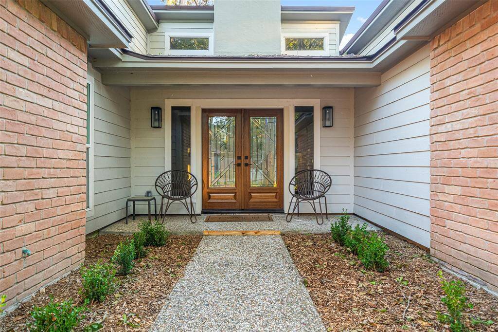 This inviting entrance features a modern double door with glass panels, flanked by stylish wall lights. The path is bordered by neatly landscaped plants, offering a welcoming atmosphere.