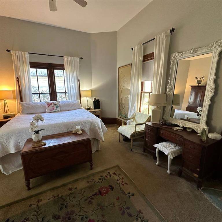 This is a cozy, well-lit primary bedroom featuring a ceiling fan, large windows with garden views, offering a comfortable and classic living space with a door leading to the porch perfect for morning coffee.