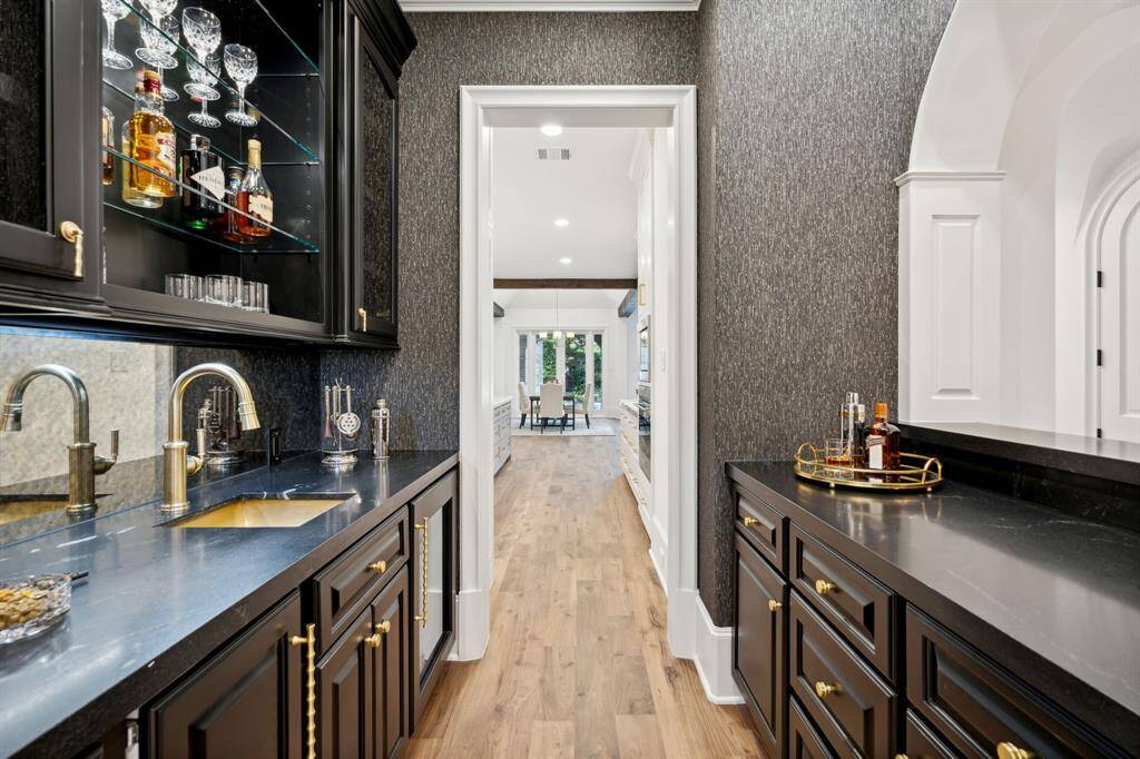 [Wet Bar] A stylish wet bar with black cabinetry, gold hardware, and a black countertop.  The area is equipped with a sink, glass shelves, antique mirrored backsplash and beautiful designer wallpaper.