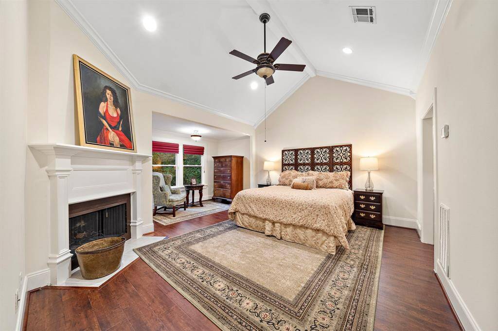 Vaulted ceilings and rich hardwood floors create a sense of elegance in this spacious primary bedroom, with a fireplace serving as a captivating focal point for the room’s refined design.