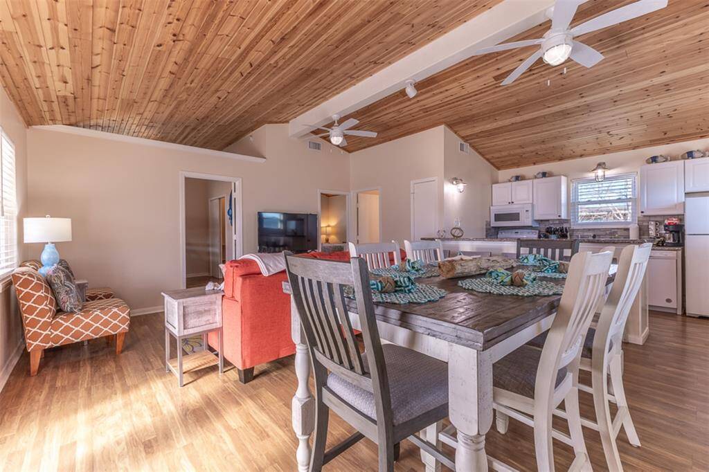 This inviting open-concept space features a cozy living area with a rustic dining table with seating for six, and a modern kitchen with white cabinets and stainless appliances. The wood-paneled ceiling and ample natural light create a warm, welcoming atmosphere.