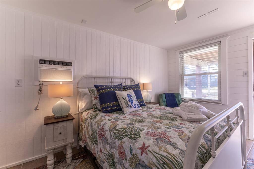 This cozy bedroom features white wood-paneled walls and a metal bed frame with floral bedding. It includes a window A/C unit, ceiling fan, and a side table with a lamp, creating a bright and inviting atmosphere.