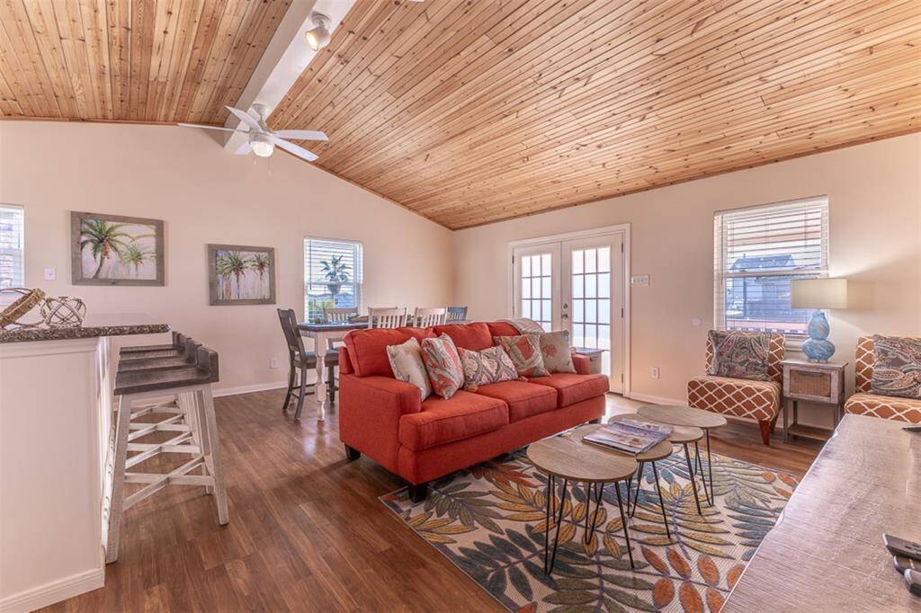 This inviting living space features a warm wooden ceiling, bright natural light from multiple windows, and cozy furnishings. The open layout includes a vibrant red sofa, stylish patterned chairs, and a dining area, creating a welcoming atmosphere perfect for relaxation and entertaining.