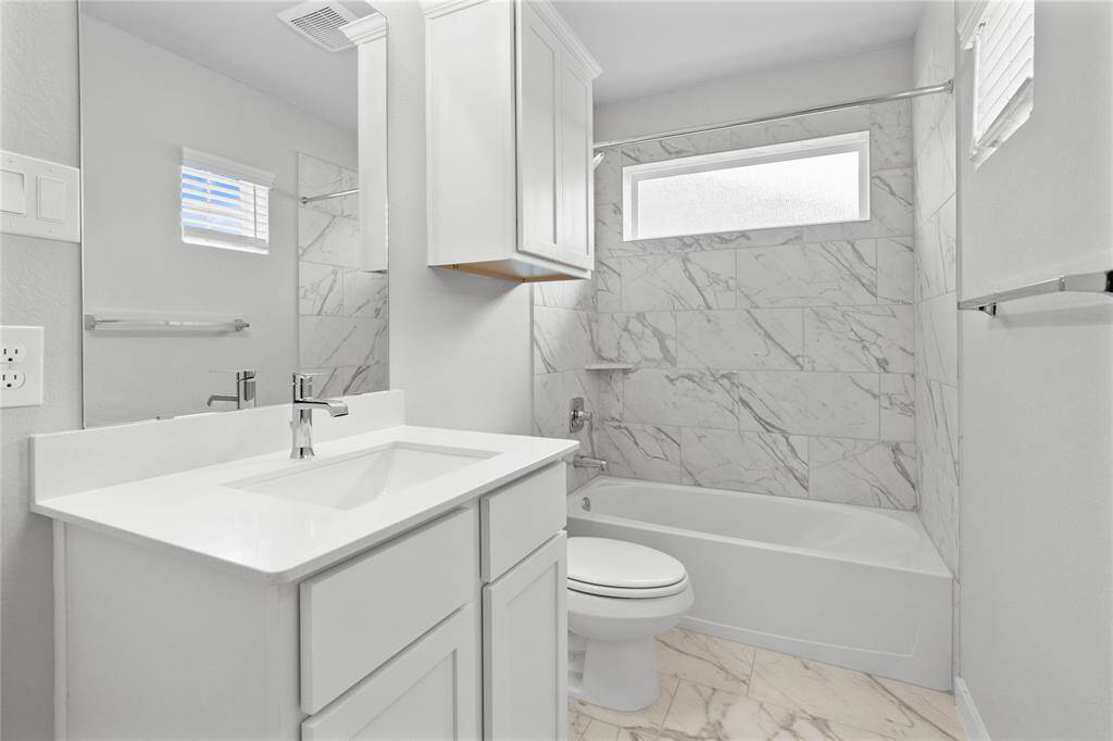 Secondary bath features tile flooring, bath/shower combo with tile surround, stained wood cabinets, beautiful light countertops, mirror, dark, sleek fixtures and modern finishes.