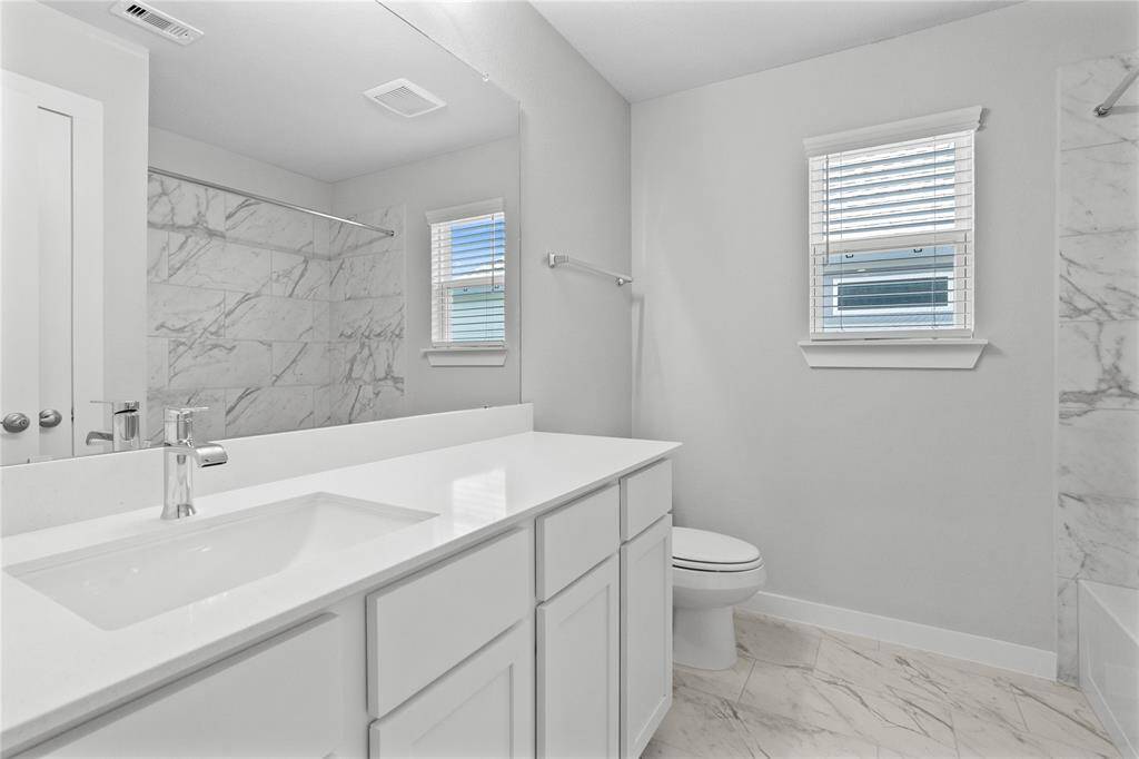 Secondary bath features tile flooring, bath/shower combo with tile surround, stained wood cabinets, beautiful light countertops, mirror, dark, sleek fixtures and modern finishes.