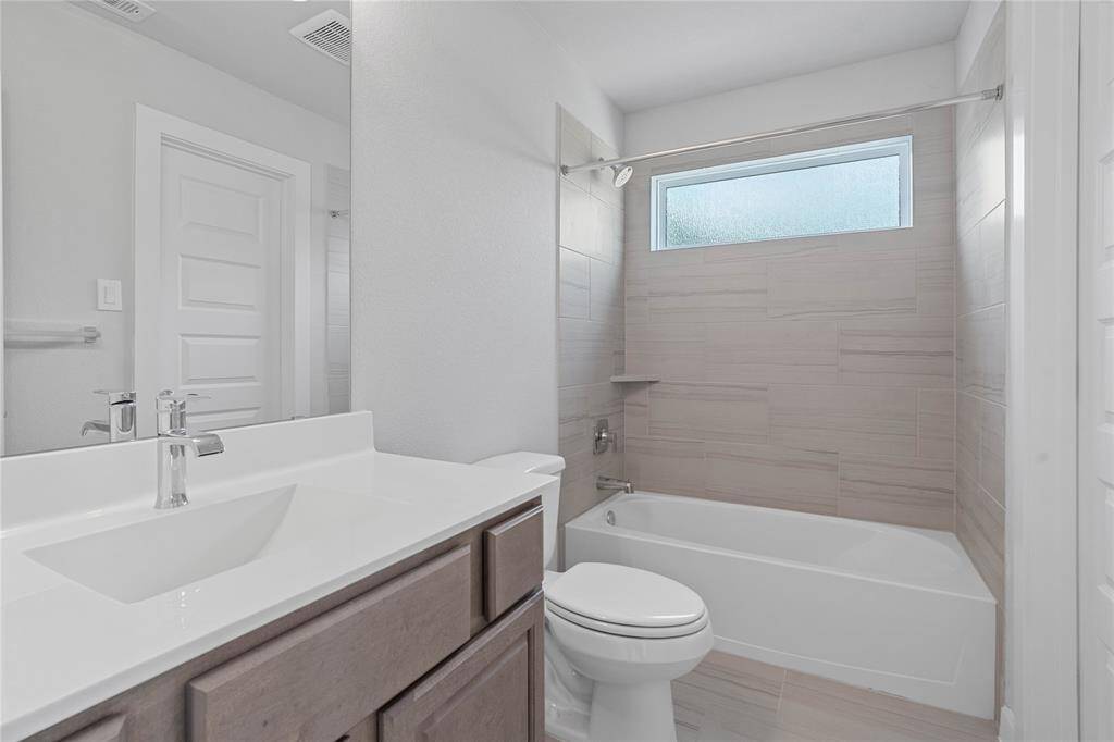Secondary bath features tile flooring, bath/shower combo with tile surround, stained wood cabinets, beautiful light countertops, mirror, dark, sleek fixtures and modern finishes.